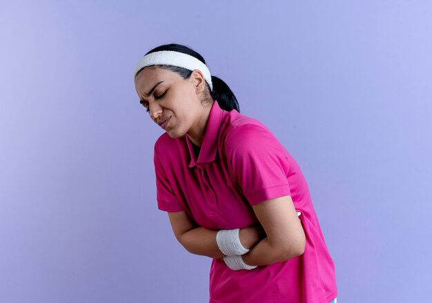 Young aching caucasian sporty woman wearing headband and wristbands holds belly on purple  with copy space