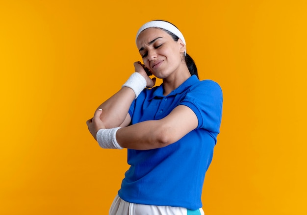 Young aching caucasian sporty woman wearing headband and wristbands holds elbow on orange  with copy space