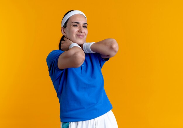 Young aching caucasian sporty woman wearing headband and wristbands holds neck looking at camera on orange  with copy space