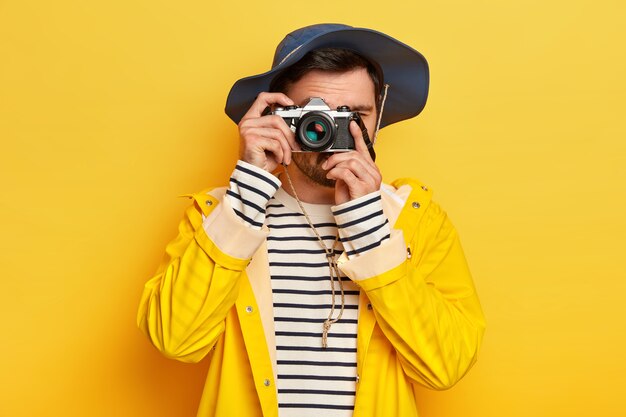 젊은 활성 남성 여행자는 비오는 날 동안 여행으로 모자, 비옷을 입은 레트로 카메라로 사진을 찍고 노란색 벽에 포즈를 취합니다.