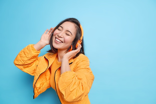 Young adorable Asian woman keeps eyes closed smiles broadly has pleased expression listens music via headphones enjoys favorite song dressed in fashionable clothes models against blue wall