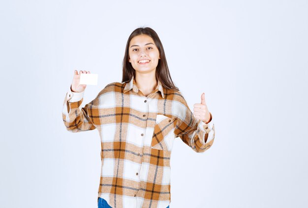 Young adorable girl showing business card and giving thumbs up on white wall. 