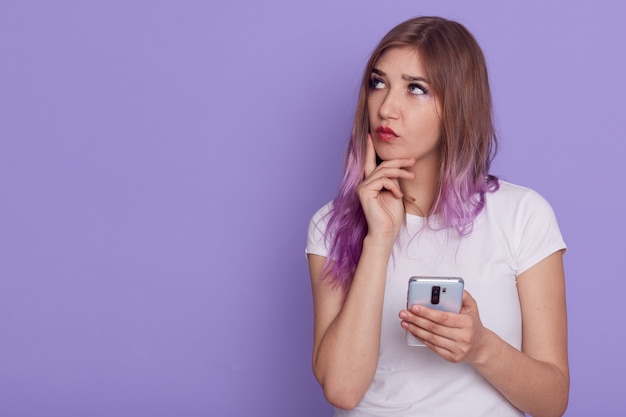 Young adorable thoughtful woman holding smart phone in hands, looking away with pensive expression, keeping finger on cheek, thinks, copy space, posing isolated over purple wall.