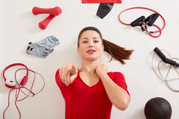 Free photo young adorable woman holding points at camera with her index finger while laying on white surface, girl with ponytail dresses red clothing