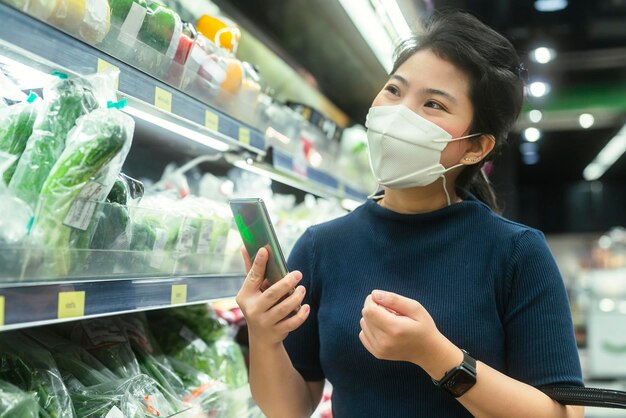 Молодая взрослая азиатская женщина в защитной маске для лица выбирает свежий продукт или потребительский товар в продуктовом магазине универмага с осторожностью и веселым новым нормальным образом жизни при покупках