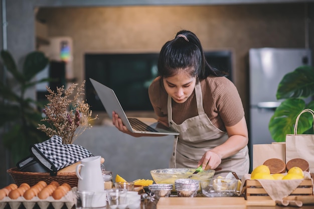 제빵 방법을 위해 노트북을 보고 있는 집 부엌에서 집에서 만든 빵집을 준비하는 젊은 성인 아시아 여성