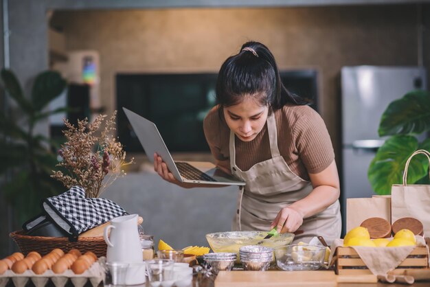 Young adult asian woman preparing homemade bakery in a kitchen at home looking on laptop for baking methods