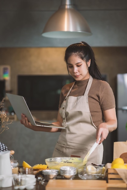 Молодая взрослая азиатка готовит домашнюю выпечку на кухне дома, глядя на ноутбук в поисках способов выпечки