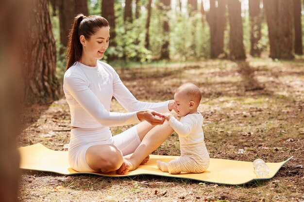 Young adult attractive mother sitting on karemat with crossed legs, holding hands her little infant child, spending time together in forest, healthy lifestyle.