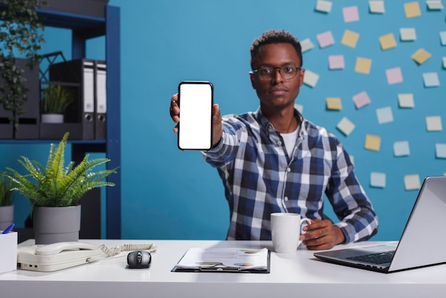 カメラに白い画面の分離されたテンプレートを表示しながら、会社のモダンなワークスペースオフィスに座っている若い大人のビジネスパーソン。モックアップディスプレイ付きの電話デバイスをカメラに向けるプロジェクトマネージャー。