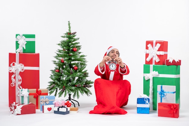 Young adult celebrate christmas holiday sitting in the ground and showing clock near gifts and decorated xmas tree