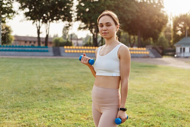 Young adult confident female wearing sporty white top and beige leggins standing in stadium with dumbbells in hands, looking directly at camera, work out outdoor.