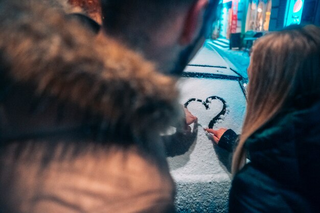 Young adult couple draws a heart on snow covered car