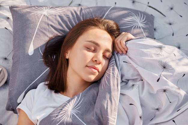 Молодая взрослая темноволосая женщина с закрытыми глазами лежит на мягкой подушке под одеялом и отдыхает