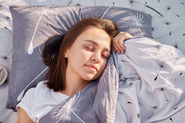 Young adult dark haired female with closed eyes lying on soft pillow under blanket and resting