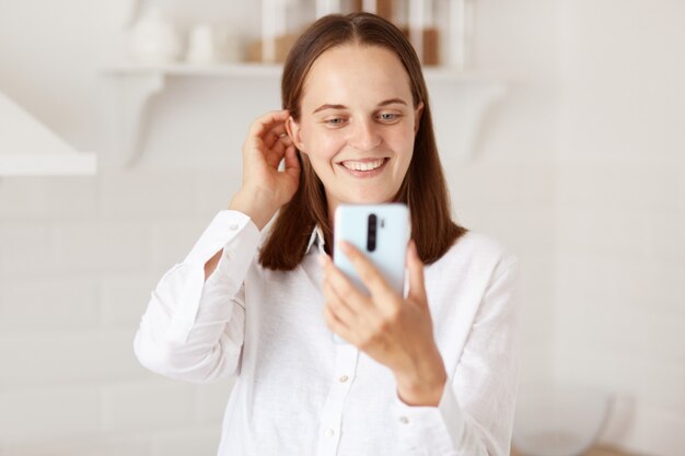 Young adult dark haired happy woman using smartphone for making selfie, posing in the kitchen at home, having pleasant conversation via video call and smiling.