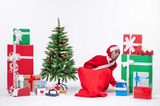 Young adult dressed as Santa claus with gifts and decorated Christmas tree sitting in the ground looking for something