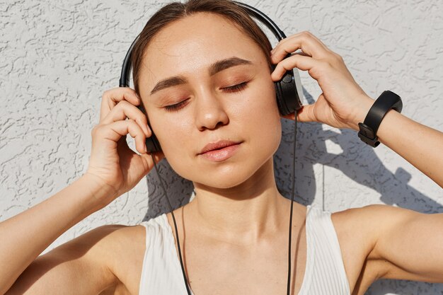 Young adult female with dark hair wearing white top, keeping eyes closed, touching headphones with palms, enjoying music after work out, healthy lifestyle.