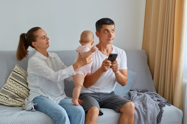 Young adult pensive man with smart phone in hands, looking away, thinks, wife with baby daughter trying to attract dad's attention, people sitting on sofa in living room.