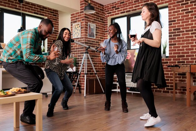 Молодые взрослые люди наслаждаются диско-музыкой дома во время празднования годовщины дружбы. Многонациональная группа закрытых друзей танцует вместе в гостиной на винной вечеринке, развлекаясь.