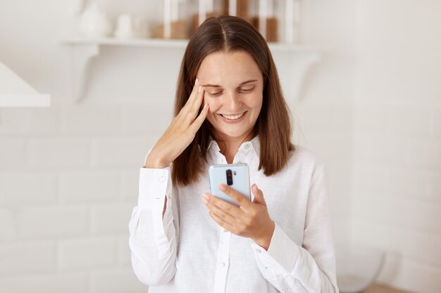 Young adult pretty happy woman using smartphone, standing with cell phone in light kitchen, keeping fingers on her temples, smiling, looking at mobile display.