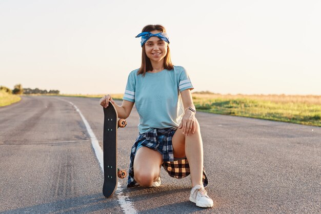 アスファルト道路で屋外にしゃがみ、スケートボードを持って、乗って休憩し、幸せな表情でカメラを見て、気持ちの良い外観を持つ若い大人の笑顔の女性。