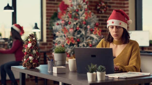 Молодой человек в шляпе Санты и работает на ноутбуке, отправляет электронное письмо с отчетом в празднично оформленном офисе. Сотрудник с помощью компьютера во время рождественских праздников. Съемка со штатива.