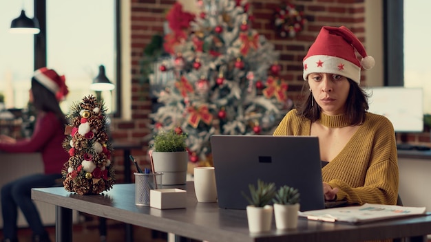 Free photo young adult wearing santa hat and working on laptop, sending email with report in festive decorated office. employee using computer during christmas holiday season. tripod shot.