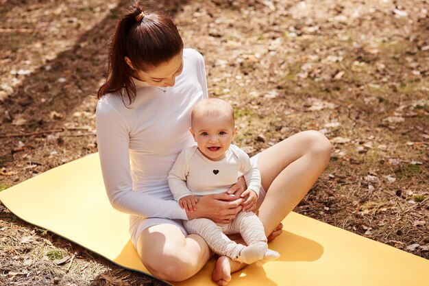 Young adult woman with dark hair wearing white clothing sitting on karemat and holding infant baby