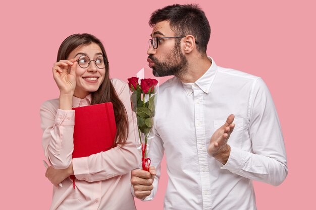 Young affectionate man gives bouquet of red roses to girlfriend, folds lips for kiss