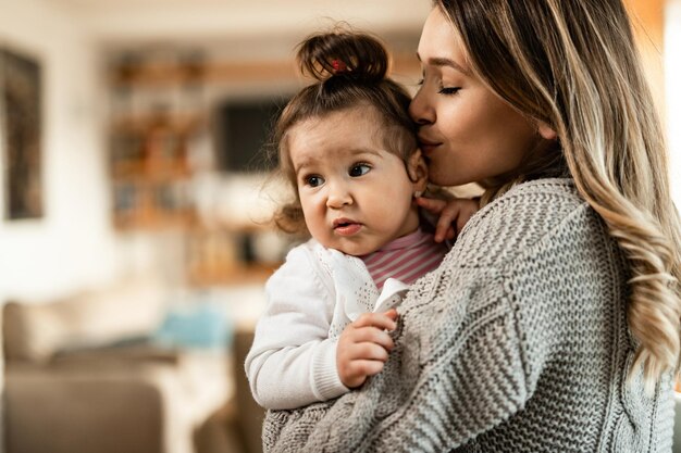 Young affectionate mother kissing her little girl while spending time with her at home