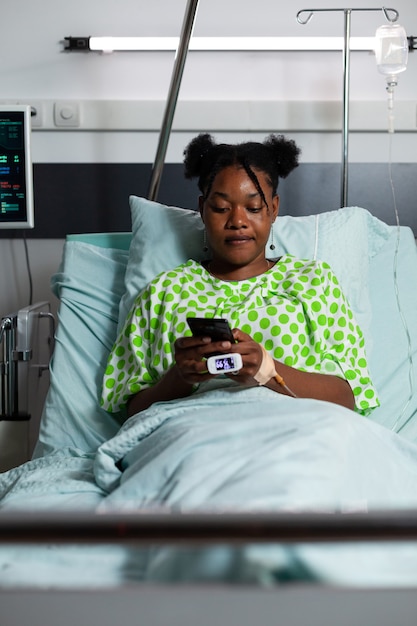 ウェブサーフィンとコミュニケーションのためにスマートフォンを使用して病棟のベッドに座っている若いアフリカ系アメリカ人の大人。オンラインガジェットを持っている間薬と相談を待っているティーンエイジャーの患者