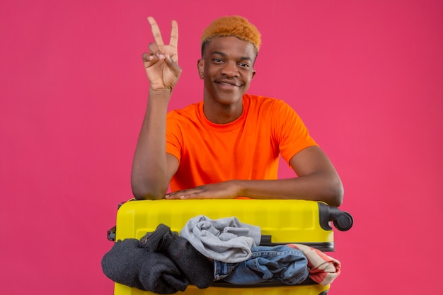 Молодой афро-американский мальчик в оранжевой футболке с дорожным чемоданом, полным одежды, смотрит в камеру оптимистично и весело улыбается, показывая номер два или знак победы на розовом фоне