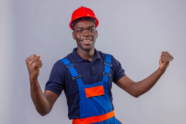 Young african american builder man wearing construction uniform and safety helmet clenching fists smiling standing with happy face celebrating victory winner