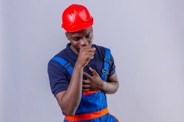 Молодой афро-американский строитель человек в строительной форме и защитном шлеме чувствует себя плохо и кашляет, стоя на изолированном белом