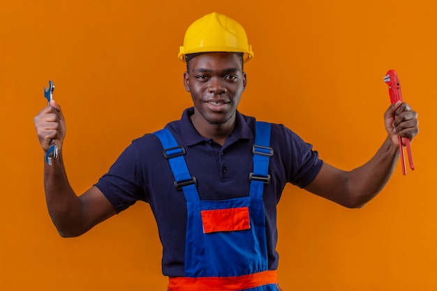 Giovane costruttore afroamericano che indossa l'uniforme da costruzione e casco di sicurezza che tiene le chiavi regolabili nelle mani alzate con la faccia felice che sta sull'arancia