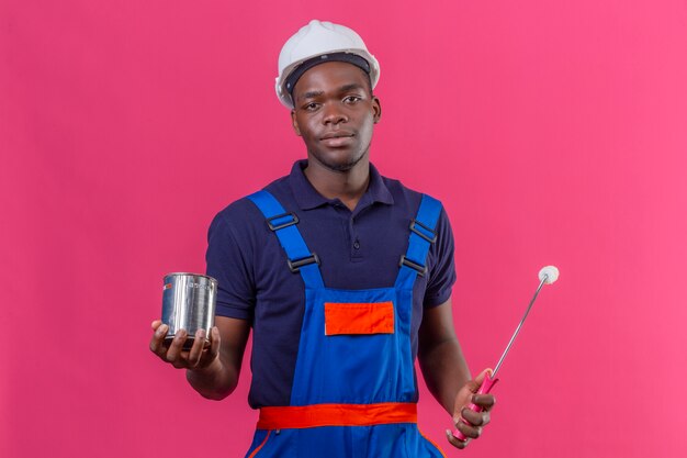 Молодой афро-американский строитель в строительной форме и защитном шлеме, держащий банку с краской и валик с уверенным серьезным выражением лица, стоящий на розовом