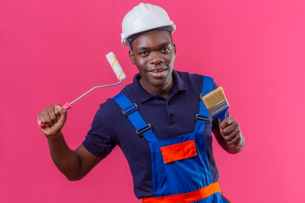 페인트 롤러와 브러시를 들고 건설 유니폼과 안전 헬멧을 착용하는 젊은 아프리카 계 미국인 작성기 남자는 분홍색에 유쾌하게 서