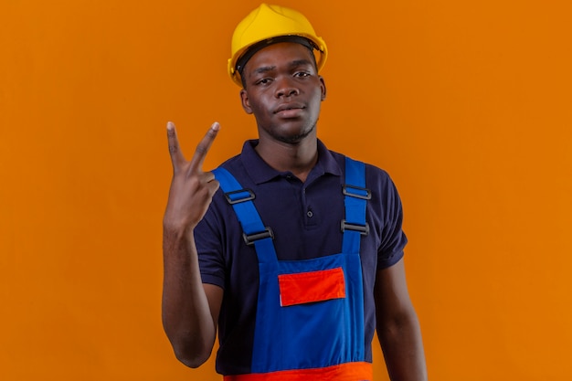 Молодой афро-американский строитель в строительной форме и защитном шлеме выглядит уверенно, показывая победу поет, стоя на оранжевом
