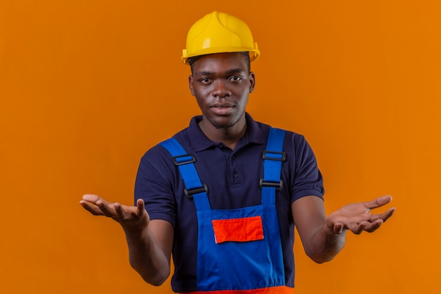 격리 된 오렌지에 질문으로 손과 표정으로 혼란스러운 제스처를 만드는 건설 유니폼과 안전 헬멧을 착용하는 젊은 아프리카 계 미국인 작성기 남자