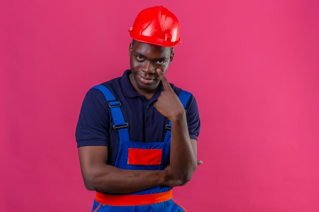 Young african american builder man wearing construction uniform and safety helmet pointing to the eye watching you gesture suspicious expression standing on isolated pink