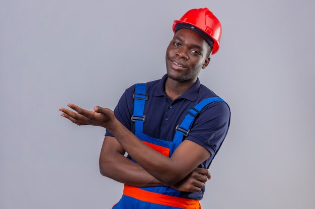 Young african american builder man wearing construction uniform and safety helmet pointing with palm of hand with smile standing 