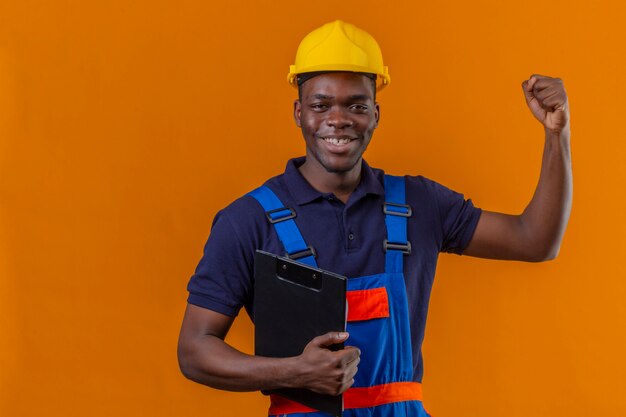 Молодой афро-американский строитель мужчина в строительной форме и защитном шлеме, стоящий с буфером обмена, поднимая руку, сжимая кулак, улыбаясь, стоя с счастливым лицом, празднуя победу