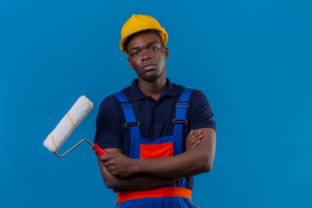 Young african american builder man wearing construction uniform and safety helmet standing with crossed arms on chest holding paint roller with frowning face displeased on