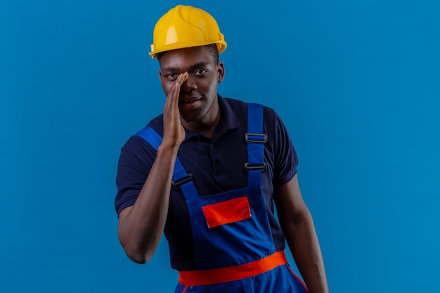 격리 된 파란색에 비밀을 말하고 입 근처 손으로 서 건설 유니폼 및 안전 헬멧을 착용하는 젊은 아프리카 계 미국인 작성기 남자