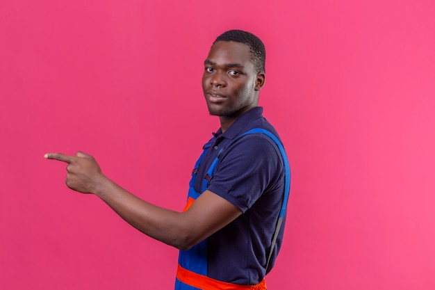 Young african american builder man wearing construction uniform standing sideways pointing to something on isolated pink