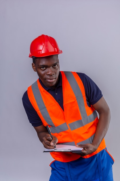 Young african american builder man wearing construction vest and safety helmet holding clipboard writing something with confident smile standing 