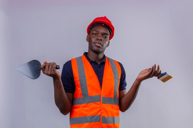 Young african american builder man wearing construction vest and safety helmet holding putty knife and paint brush standing with confused expression with arms and hands raised 