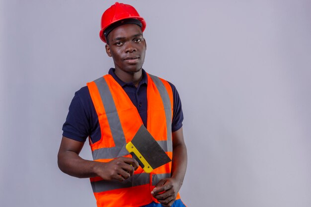 Young african american builder man wearing construction vest and safety helmet holding putty knife with serious face standing 