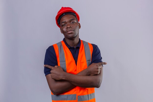 Молодой афро-американский строитель мужчина в строительном жилете и защитном шлеме держит руки, скрещенные, указывая пальцами с уверенным взглядом стоя
